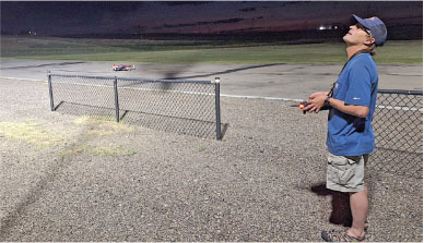 Man in blue shirt controlling a remote plane at dusk near a chain-link fence.