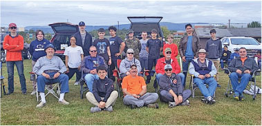 Group of people posing outdoors on grass, some seated, cars in the background.