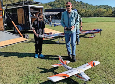 Two people with controllers near remote-controlled planes on grass.