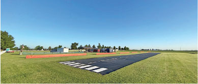 Runway on grassy field under clear blue sky. Buildings in the background.