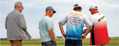 Four men standing in a grassy field, one wearing a "VOLUNTEER" shirt.