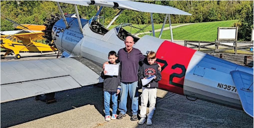 Three people standing in front of a vintage biplane; one holds a certificate.