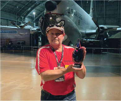 Man in red shirt holding an award, with a space shuttle in the background.