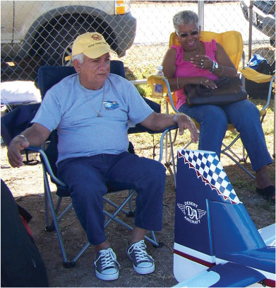 Elderly man and woman sitting on chairs outdoors by a model aircraft.