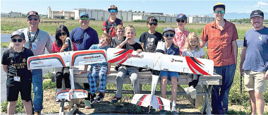 Group of people outdoors with remote-controlled planes on a bench.