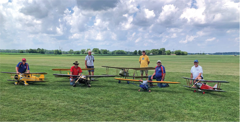 Five men with model planes on a grassy field under a partly cloudy sky.