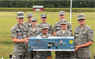 Seven cadets in uniform holding a HobbyZone Aeroscout S 2 1.1m model airplane box outdoors.
