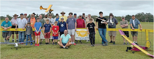 Group of people with model airplanes, smiling outdoors near a yellow fence.