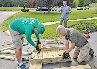 Two men assembling wooden boards with drills, a third man stands nearby on a driveway.