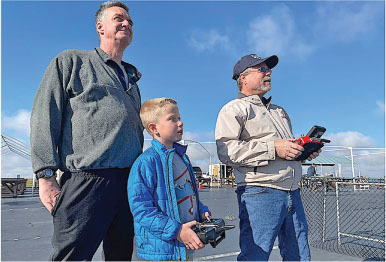 Two men and a boy control a drone outdoors, sky in the background.