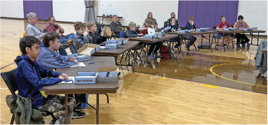 Students in a classroom setting, seated in rows at tables, with books and notes.