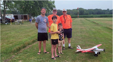 Five people standing on grass with model airplane in the foreground.