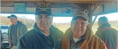 Two men smiling in a sheltered outdoor area on a sunny day, wearing jackets and caps.