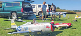 Model airplanes on grass near parked trucks, with two people standing beside them.