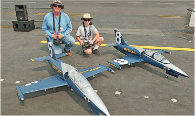 Two people kneeling with large blue model jets labeled "BREITLING" on a runway.