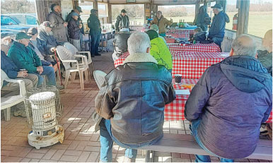 Men gathered in a shelter with red checkered tables, heater, and outdoor view.