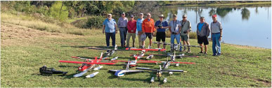 Group of people standing behind model airplanes on a grassy field by a lake.