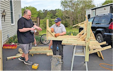 Two men assembling wooden structure on driveway with tools around.