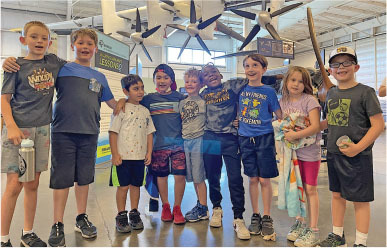 Group of eight children smiling inside, airplane model above.