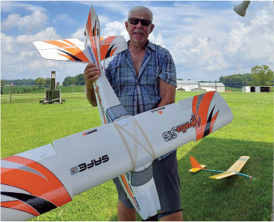 Man holding an orange and white model plane on a grassy field.