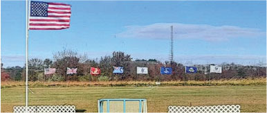 Flags on poles in a field, including a prominent U.S. flag, under a blue sky.