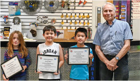 Three children hold certificates beside an older man in a workshop.