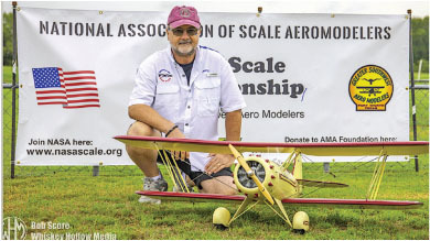 Man kneeling beside scale model plane, National Association of Scale Aeromodelers banner behind.