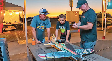 Three people assembling a model airplane on a table at sunset.