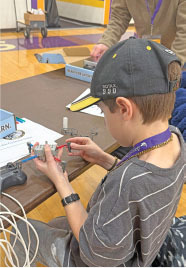 Boy assembling a small robot at a table, wearing a cap and striped shirt.