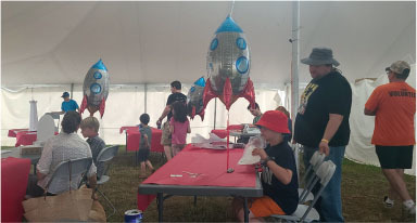 Children and adults at a tent event, with rocket balloons and red tables.