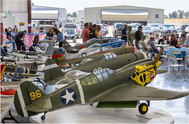 Model airplanes displayed in a hangar with people and tables in the background.