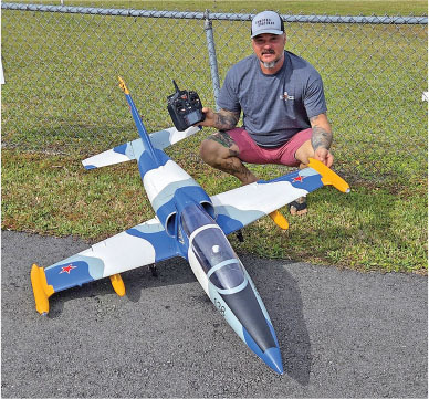 Man crouching next to large model jet with remote control on grass.