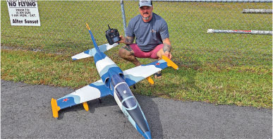 Man with remote control next to a large model jet on grass, chain link fence in background.
