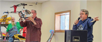 Man holding helicopter model, another speaking into a microphone in a room.