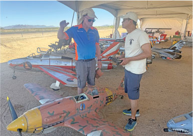 Two people conversing next to model planes under a tent in a sunny outdoor setting.