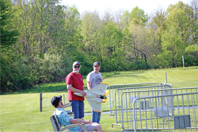 Three people by a grassy field with a model airplane, trees in the background.