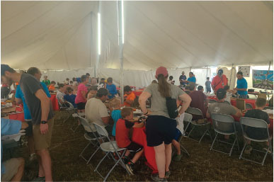 People seated and standing under a large, white tent, engaged in various activities.