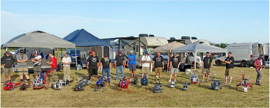 Group of people standing with model helicopters on grass, tents and RVs in background.