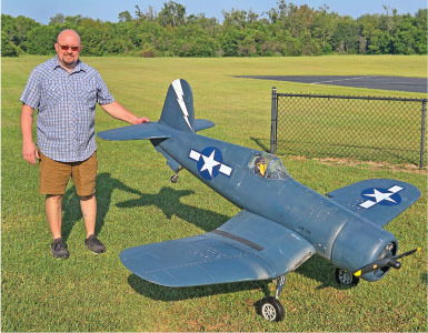 Man in shorts standing next to large blue model plane on grass.