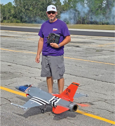 Man with remote control stands beside a model jet on an airstrip.