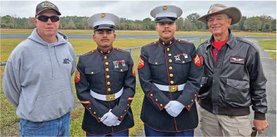 Two Marines in dress uniforms stand between two men in casual jackets and hats.