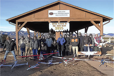 Group posing with model airplanes under a pavilion with "We're flying in the New Year 2025" banner.