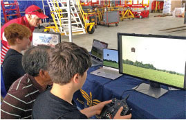 People using computers and a remote controller with a simulation on screen, in a hangar setting.