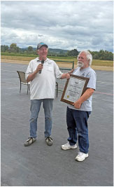 Two men standing outdoors, one holds a microphone, the other a framed certificate.