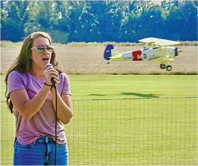 Woman singing outdoors with small plane in background.