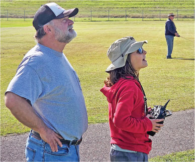 Man in cap and woman with remote control on grass field, another person in background.