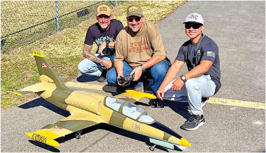 Three men kneeling on pavement with a model aircraft, smiling.