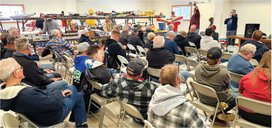 Audience seated indoors, listening to a speaker near tables of model airplanes.