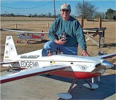Man kneels beside large remote-controlled plane, smiling, outdoors on a clear day.