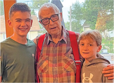 Elderly man with red straps embraces two smiling boys indoors.